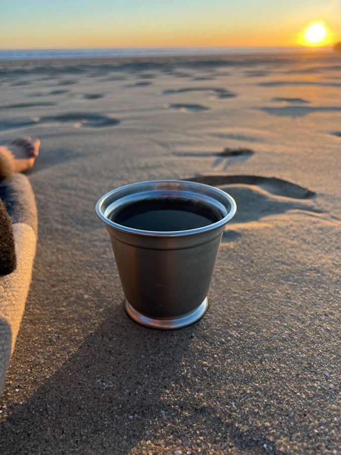 Illustration: a plain steel cup with black coffee on a blanket on the beach at sunrise.