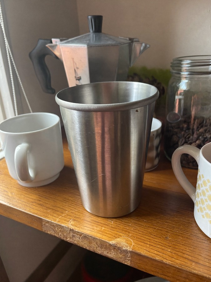 Illustration: a plain steel cup on a kitchen shelf between ceramic mugs and a moka pot.